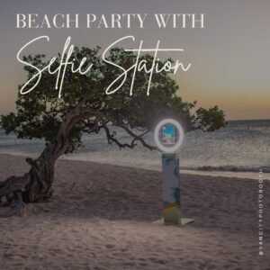 Selfie Station photo booth rental setup on a beach at sunset, ring light glowing beside a windswept tree