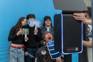 Group of students using a Selfie Station with playful photo booth props in front of a bright photo booth backdrop at a youth event.
