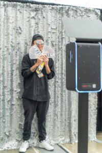 Guest poses with money-themed photo booth props in front of a sequin photo booth backdrop, captured at a Vancity Photo Booth rental event.