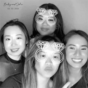 A group of four women poses together in a black-and-white GIF photo booth, some wearing fun diamond ring glasses at Becky and John’s wedding on February 3, 2024.