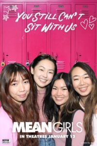 A group of four smiling women posing in front of a hot pink locker backdrop inside a themed photo booth experience for the "Mean Girls" movie release.