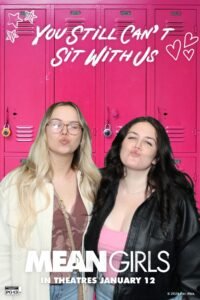 Two women pose in front of a vibrant pink high school locker backdrop inspired by Mean Girls, blowing kisses at the camera. The photo booth overlay reads “You Still Can’t Sit With Us” with hearts and stars, along with the movie title and release date.