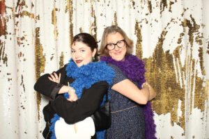 Two women pose confidently in front of a gold-and-white sequin backdrop at a photo booth, wearing colorful feather boas and playful smiles.