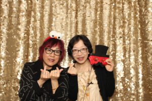 Two women pose with fun props and smiles in front of a sparkling gold sequin backdrop at a photo booth rental in Vancouver.