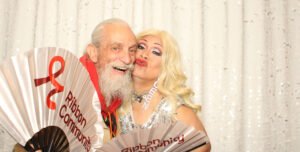 An older man and a glamorous senior drag queen smile and pose together in a photo booth, holding “Ribbon Community” fans against a sparkling sequin backdrop.