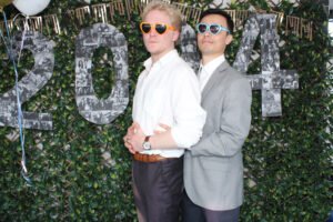 Two men wearing heart-shaped sunglasses pose playfully in front of a leafy green backdrop with large collage-style “2024” numbers at a photo booth in Vancouver.