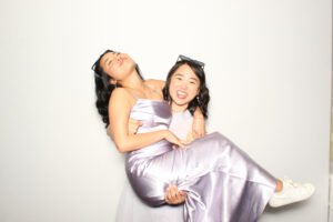 Two women in satin lavender gowns pose playfully in a photo booth, with one carrying the other while both laugh joyfully in front of a clean white backdrop.