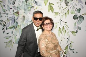 A well-dressed older couple smiles while wearing sunglasses in a floral-themed photo booth, celebrating at a formal event in Vancouver.
