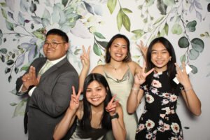 Four guests in formalwear pose playfully in front of a leafy floral photo booth backdrop inside a photo booth at a Vancouver event. Everyone is smiling and flashing peace signs.