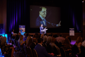 A keynote speaker in a blue suit addresses a seated audience at a formal event, with his image projected on a large screen behind him on stage.