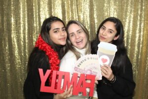 Three women pose in front of a glittering gold backdrop, holding fun photo booth props including a red “FAMILY” sign, fake money, and a cartoon coffee cup with a heart.