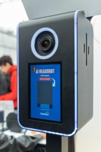 Close-up of a modern AI-powered photo booth with a built-in camera, touchscreen interface, and ring light, branded as “AI Headshot” and sponsored by Suncor at a professional event.