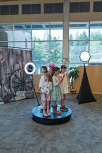Children standing on a 360 photo booth platform in front of a rustic Western backdrop during a family-friendly indoor event in Vancouver.