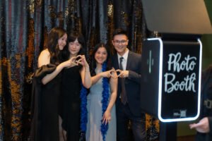 Group of young adults posing together at a Vancity Photo Booth with a sequin backdrop, making heart signs and smiling during a formal event.
