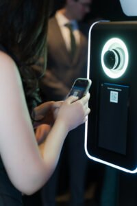 A guest scans a QR code on a modern photo booth screen using their phone at a sleek event. The booth features a glowing ring light and digital interface, designed for instant photo sharing.