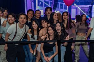 Group of young adults smiling and posing at a lively indoor event with purple lighting and balloons.