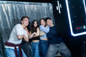 Four friends strike fun poses in front of a silver sequin backdrop at an open-air photo booth.