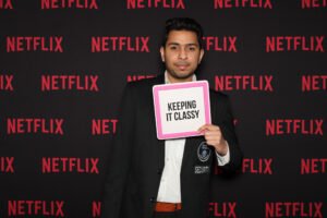 A well-dressed man poses in front of a step-and-repeat Netflix-branded backdrop, holding a sign that reads “KEEPING IT CLASSY” with a confident expression.