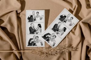 Black and white wedding photo booth strips featuring a bride and groom posing playfully with drinks and straws, laid on soft tan fabric with a dried grass accent.